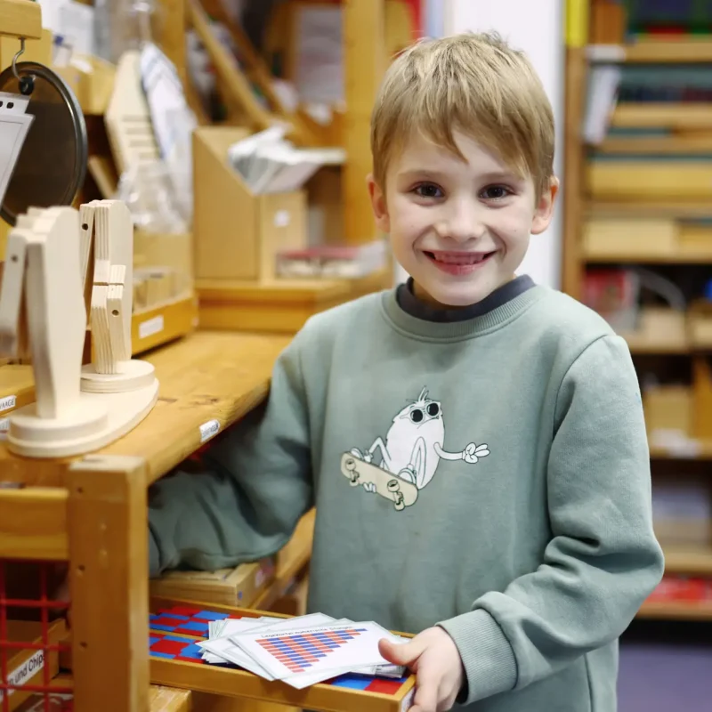 Kind zeigt stolz ein Montessori-Material und lächelt in die Kamera.