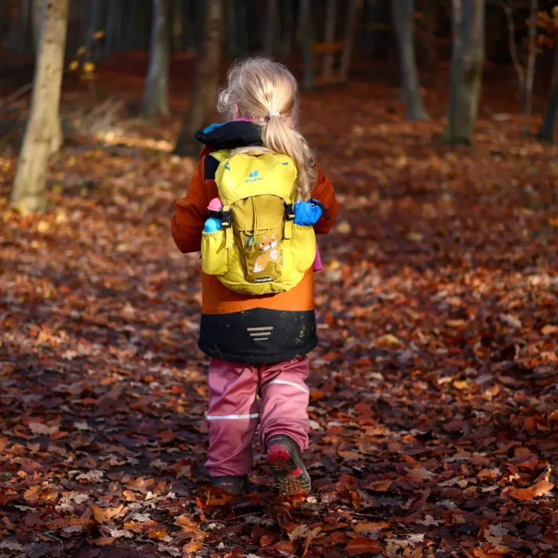 Kind mit gelbem Rucksack steht auf einem Laubboden und schaut in den Wald hinein.