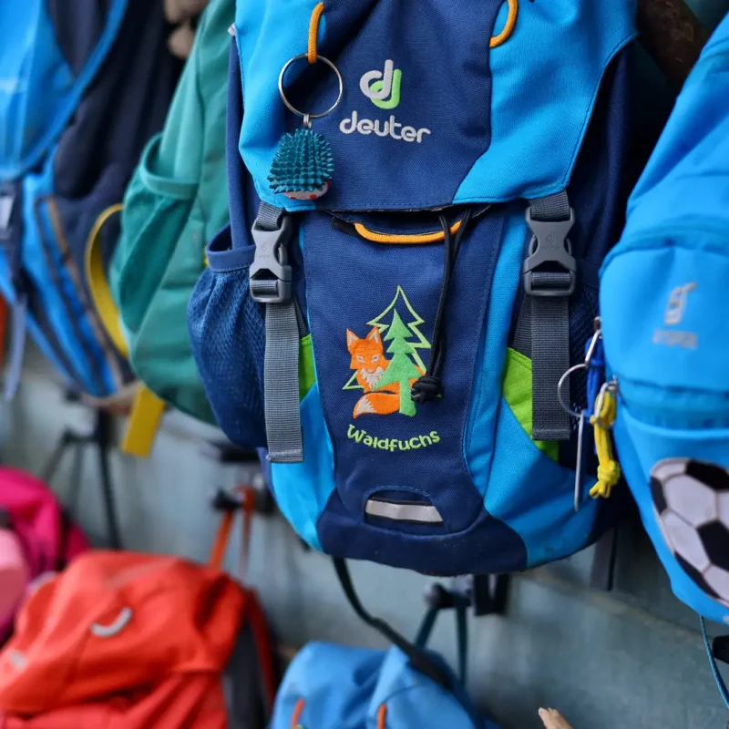 Mehrere farbige Kindergartenrucksäcke hängen an einem Holzgestell im Wald.