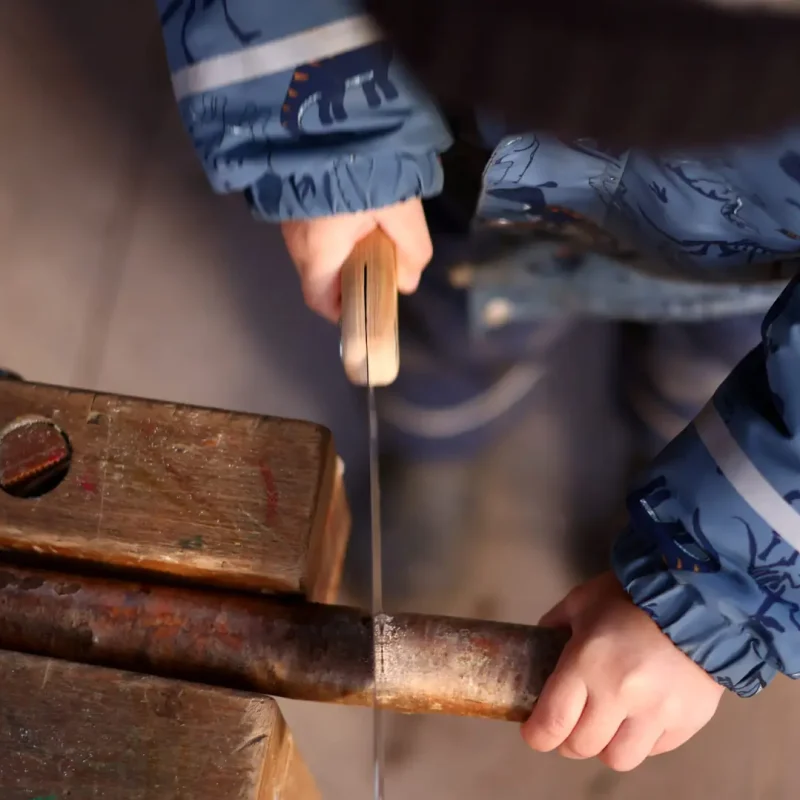 Kind arbeitet mit einem echten Werkzeug und sägt vorsichtig an einem Holzstück.