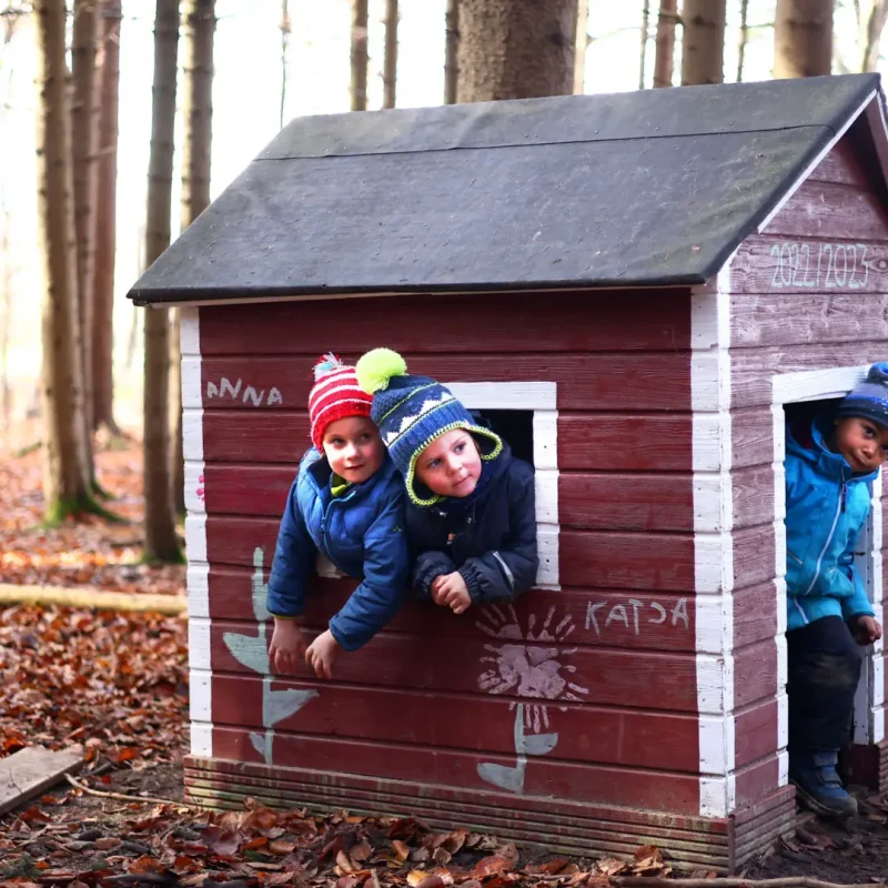 Kinder spielen im kleinen roten Holzhaus und schauen aus den Fenstern heraus.