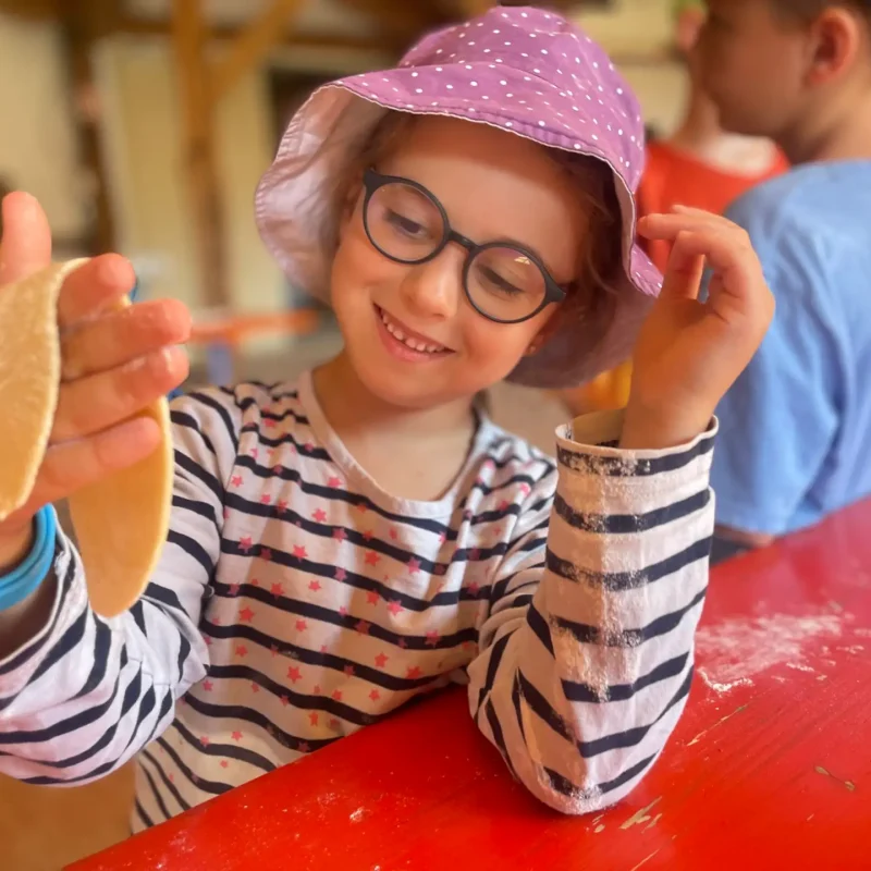 Mädchen mit Brille sitzt am Tisch, zeigt stolz ihren Teig und lacht.